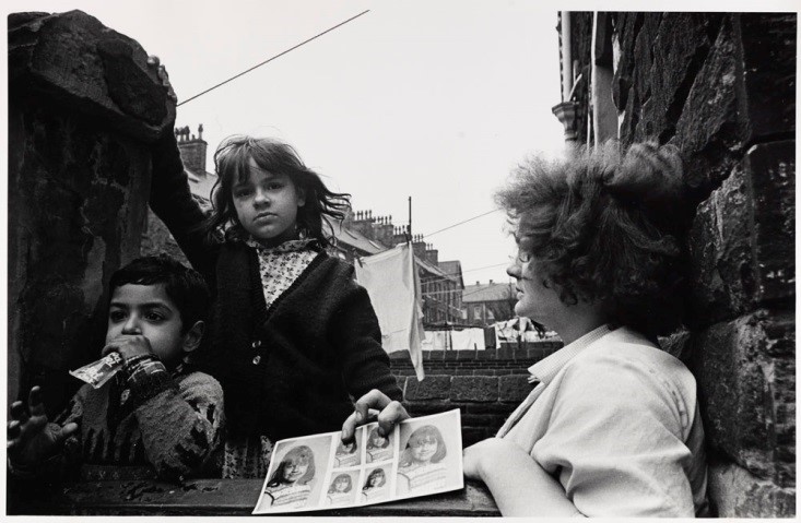 Untitled print of woman and children outside terraced housing in Bradford, showing sheet of school photographs by Nick Hedges