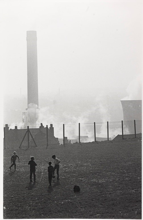 Boys playing football on waste ground; chimney and cooling tower in background, Bradford by Nick Hedges