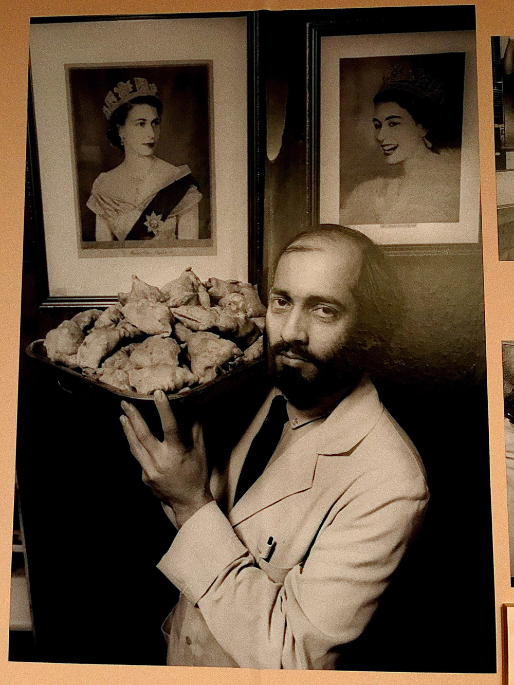 Black-and-white photograph of a man holding a plate of samosas with a portrait of the Queen in the background