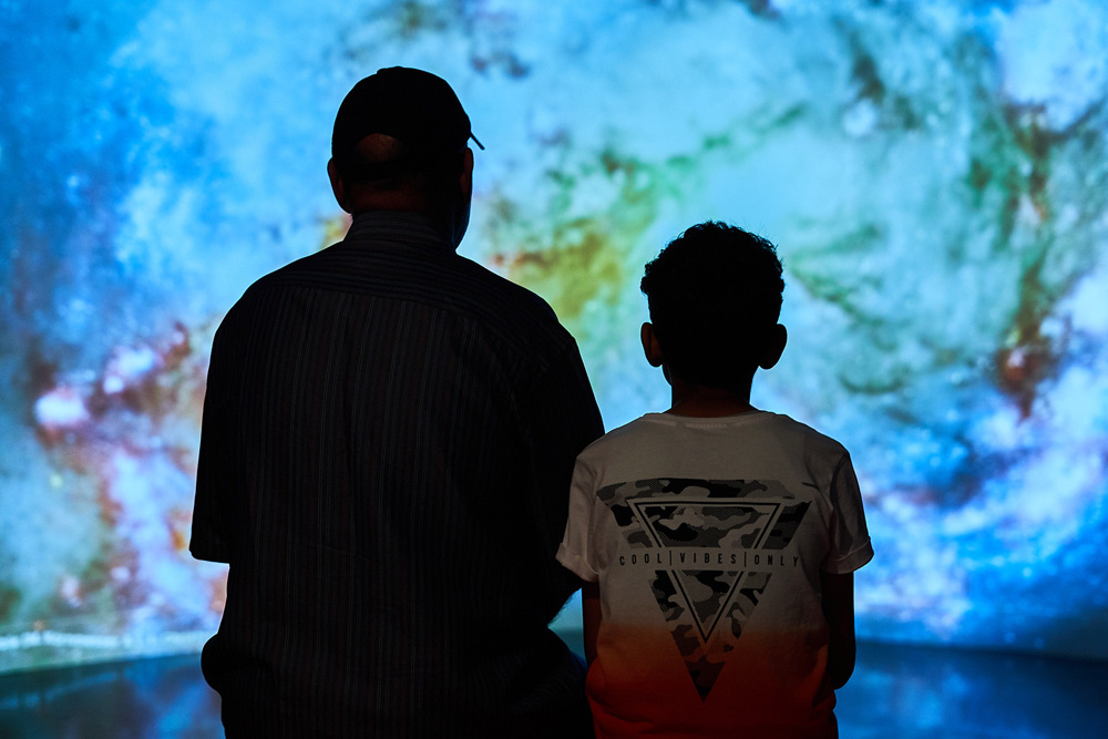 A man and a boy (in silhouette) sit in front of a large screen showing images of space