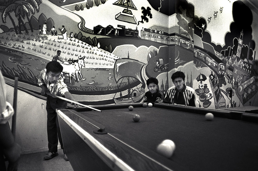 Three young men playing pool in front of a mural depicting Bangladesh and Bradford