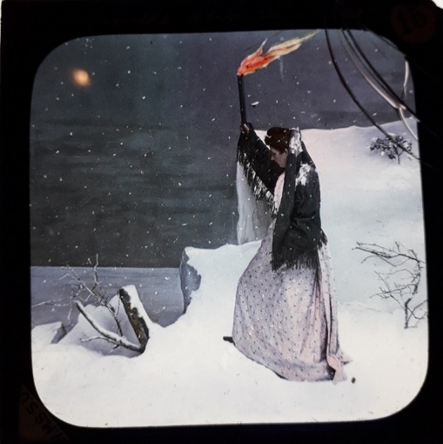 Lantern slide showing woman standing near a cliff in heavy snow, looking out at the sea, holding a lit flare