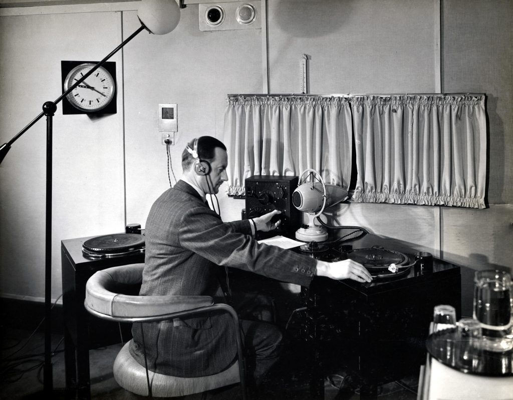 Suited man wearing a microphone sits at a desk operating radio equipment
