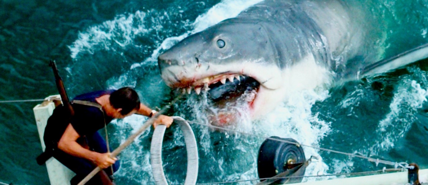 A shark with blood on its jaws dives from the water toward a man on the mast of a boat