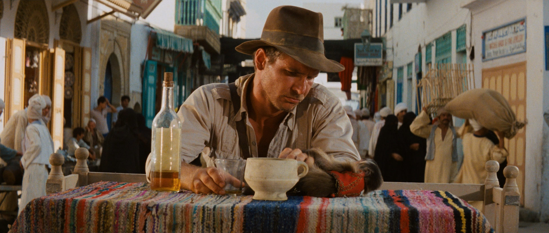 Harrison Ford sits drinking liquor at a table in a bustling middle eastern street