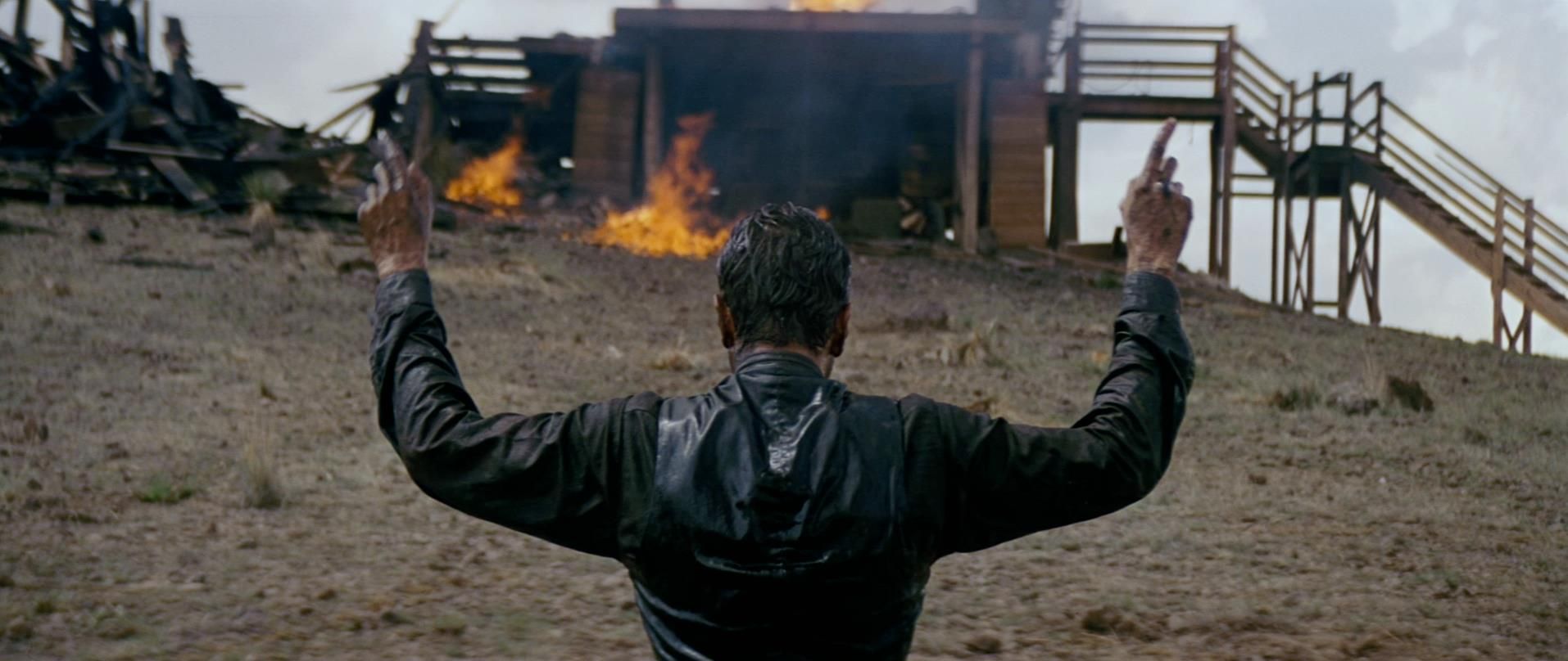 A man wearing black holds up both hands as he watches a wooden building set on fire