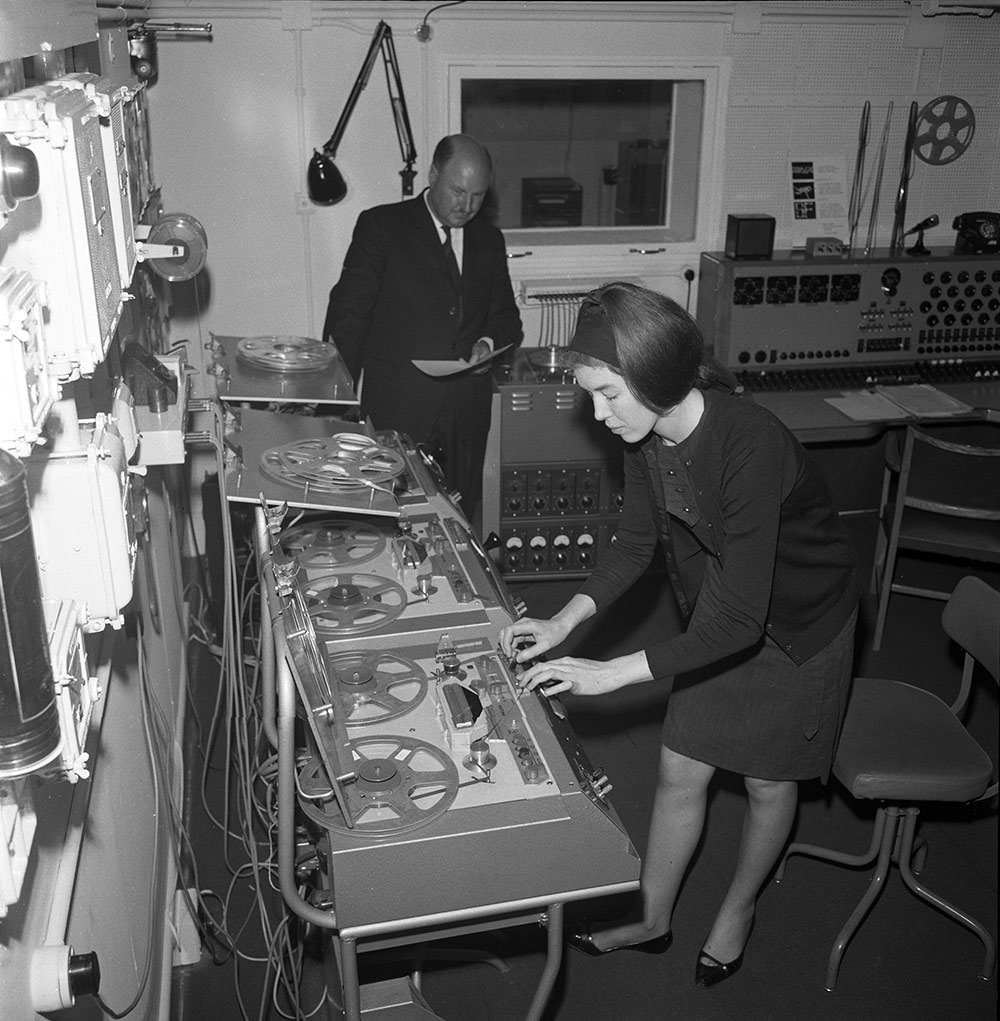 Woman adjust the controls on a tape deck while a man in a suit looks on