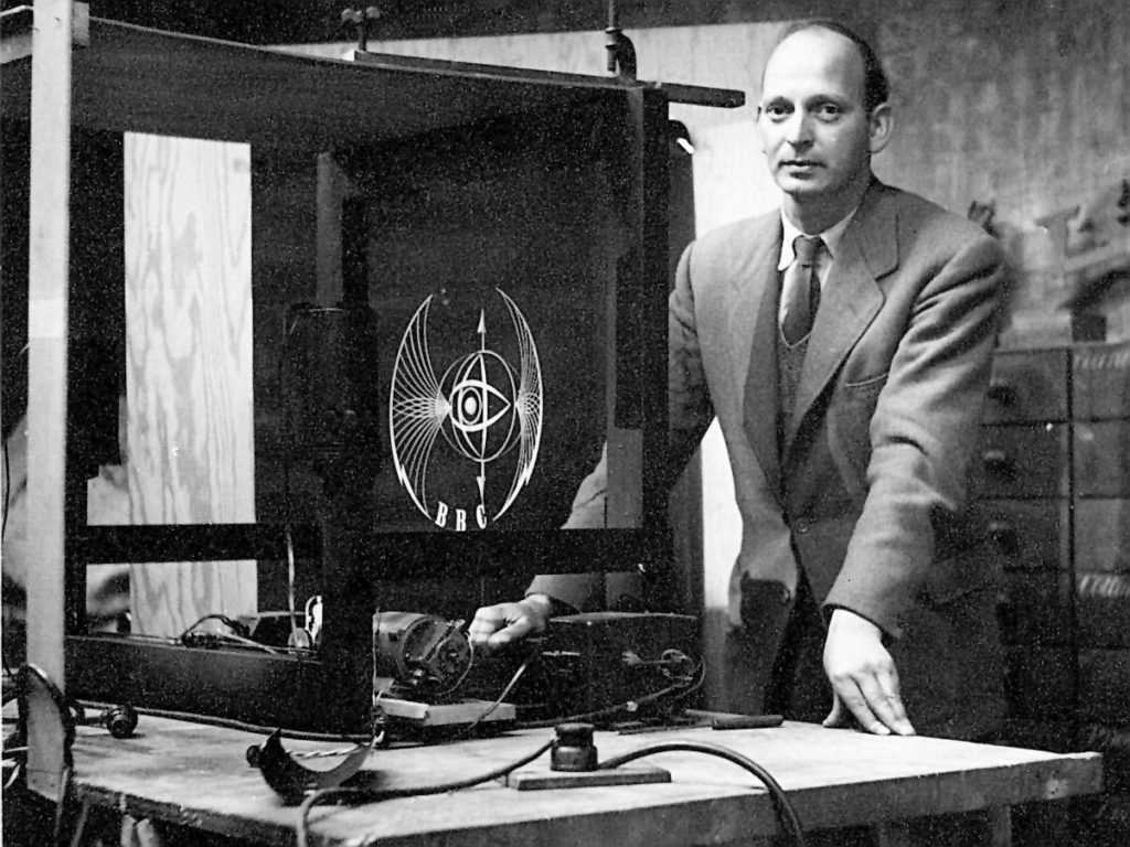 Abram Games stands by a tabletop with electrical cables and a wooden frame containing a model of his BBC emblem.