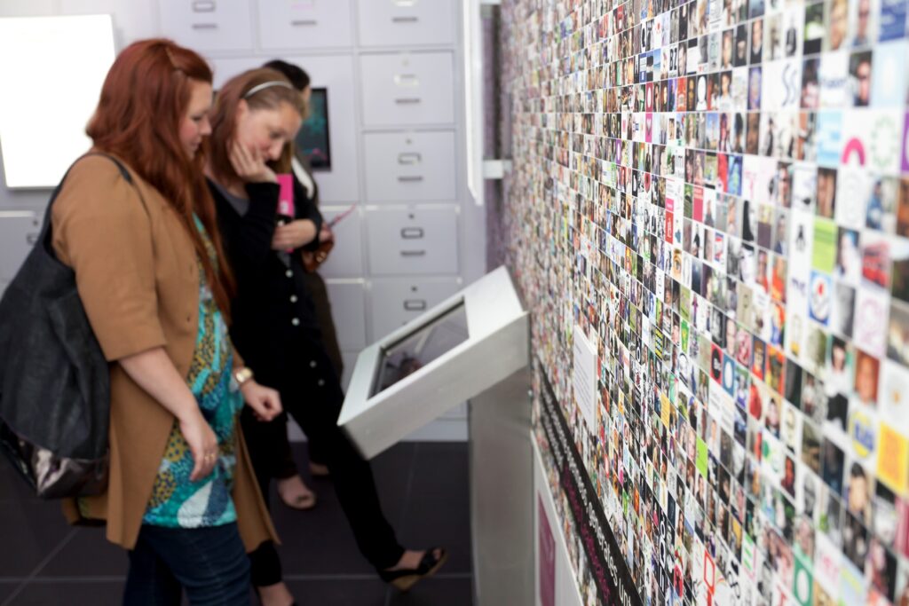 Two people looking at a screen, next to a wall covered in a photo montage