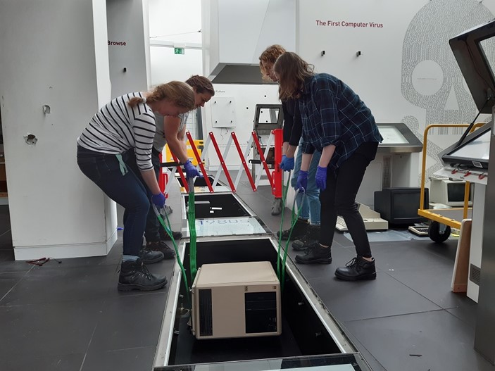 Four museum staff use straps to lift a computer up from an under-floor display.