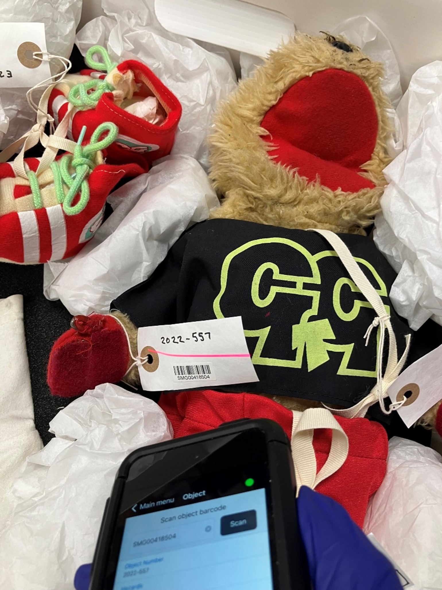 A fluffy toy gopher and his red shoes pack in tissue paper, with a gloved hand holding a smart device for cataloguing