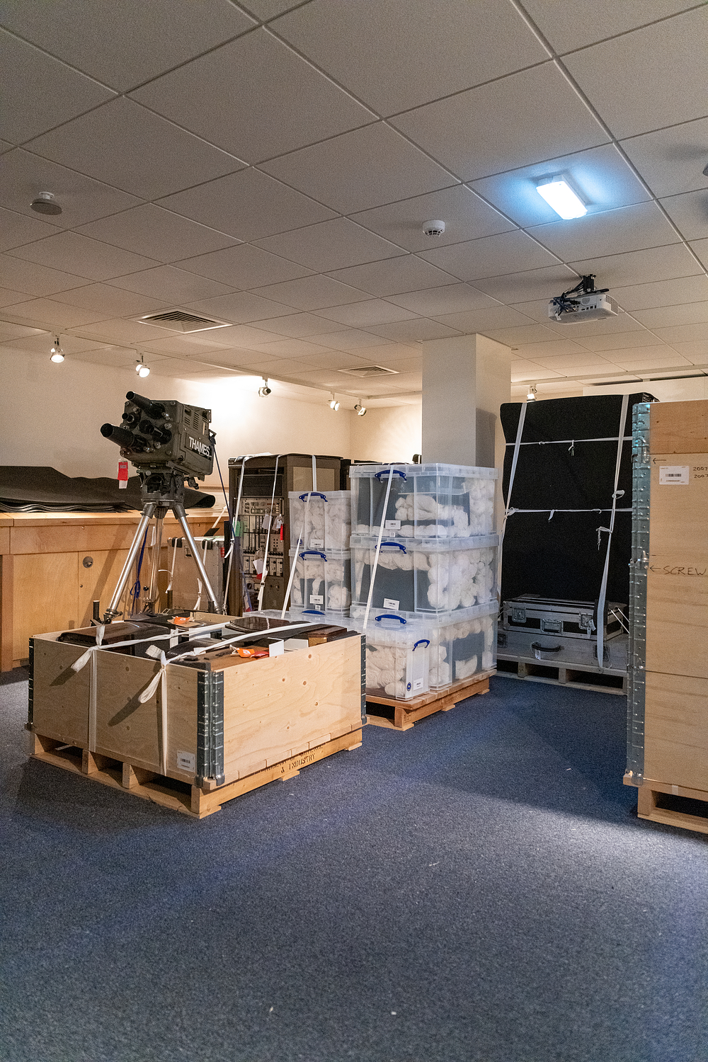 A room full of wooden and plastic crates full of museum objects, with a tv camera balance on top one of the boxes