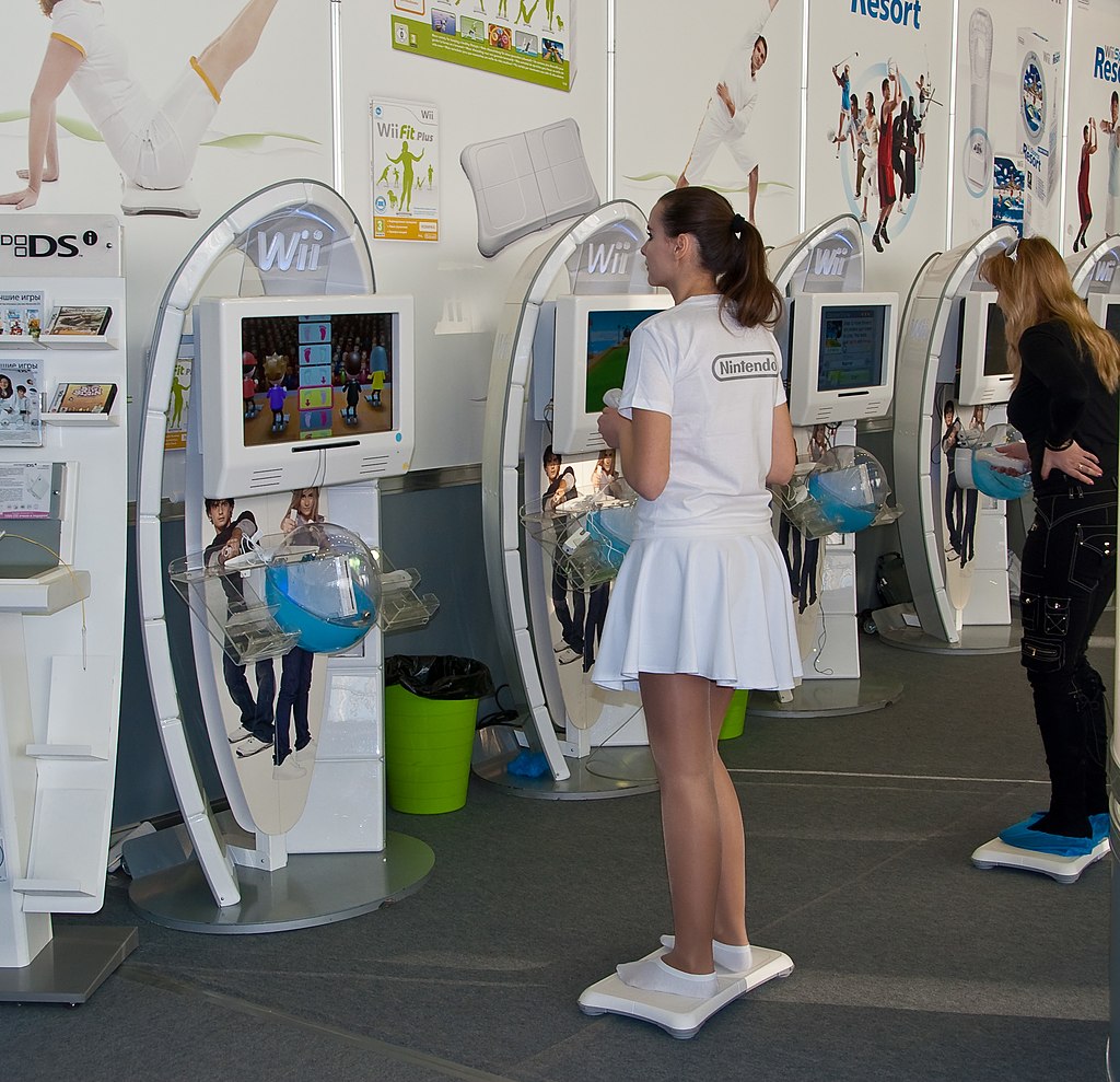 Two women stand on Wii balance boards in front of a row of screens, playing a Wii game
