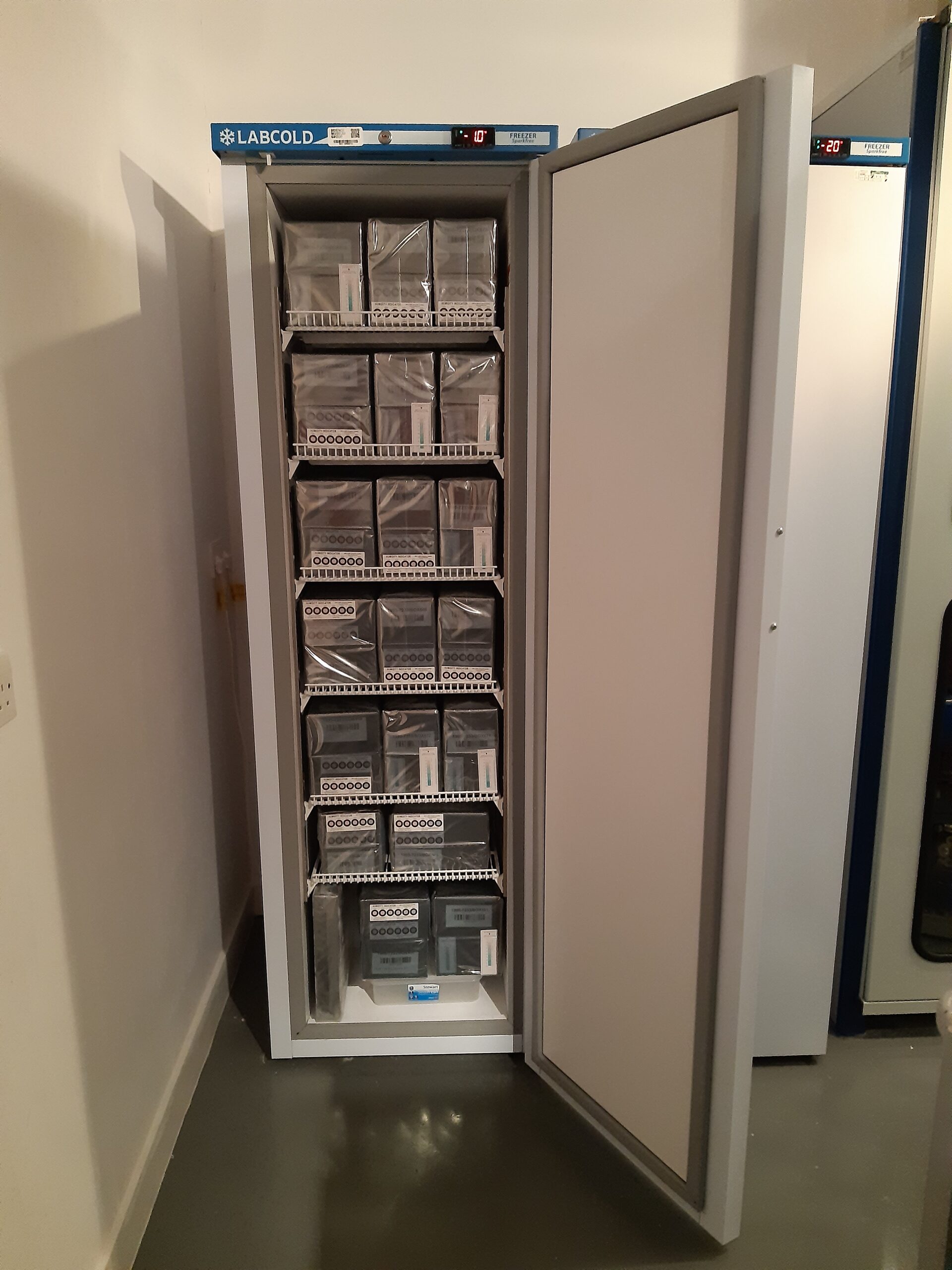 A tall thin freezer with the door open, showing shelves full of black boxes