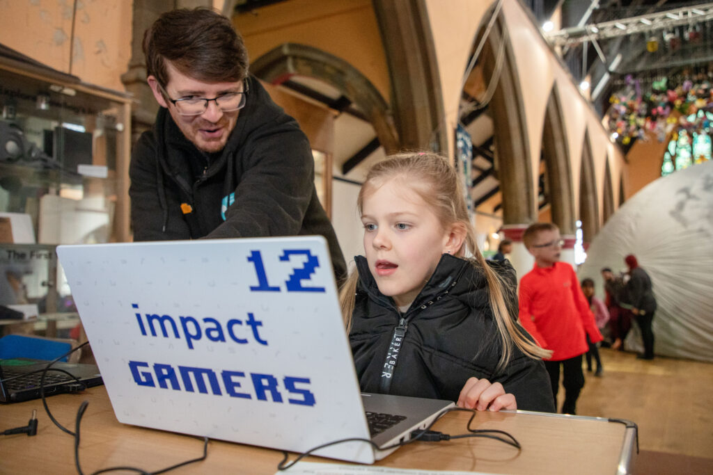 A young girl looks at a laptop with 'impact gamers' on the back, helped by an adult
