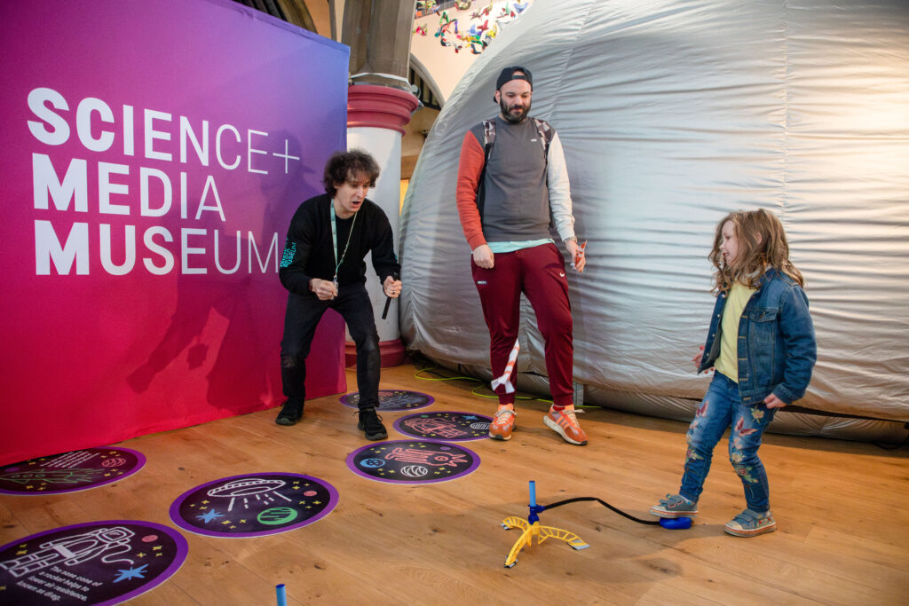 A museum explainer cheers on a child as they launch a stomp rocket, while another adult looks on