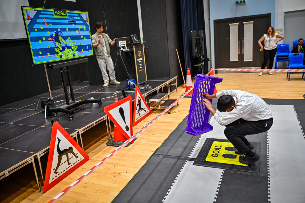 A young person playing a digital game uses a plastic crate to shield themselves while their avatar appears on a screen