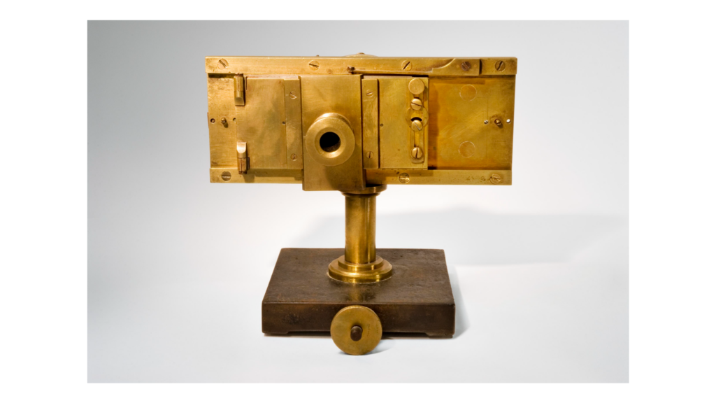 A brass-coloured early camera in a simply rectangle shape, mounted on a wooden stand.