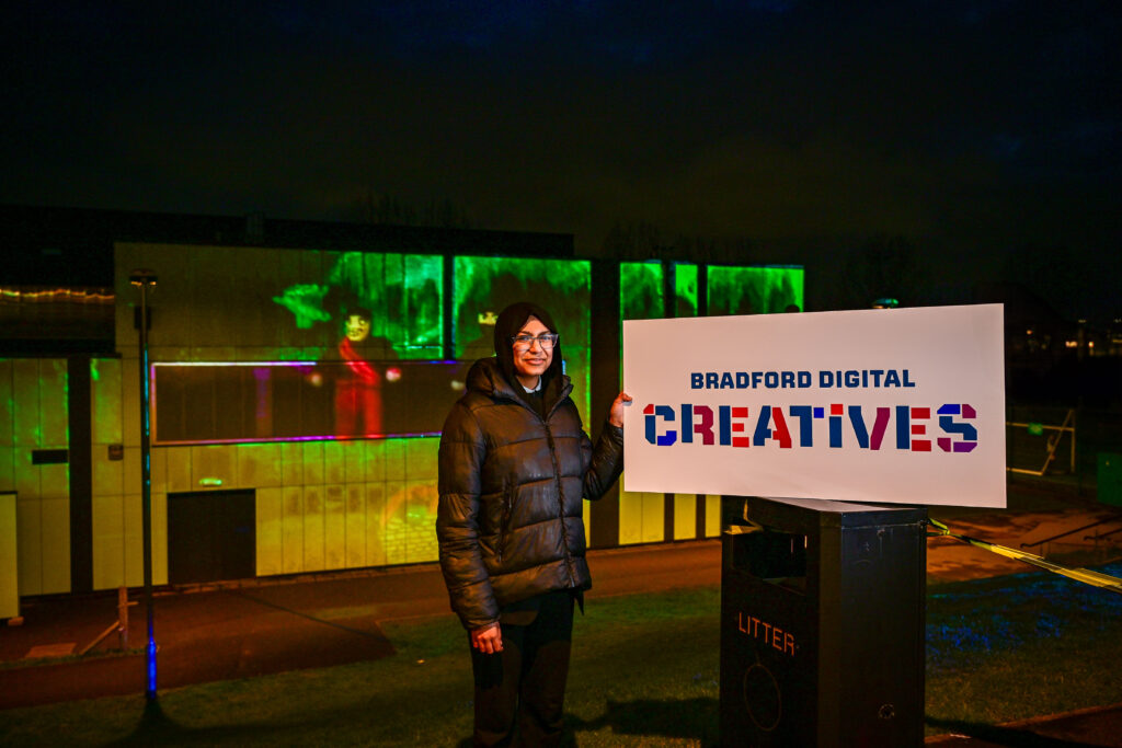 A student holding a sign reading Bradford Digital Creatives stands by a building with an animation projected onto it