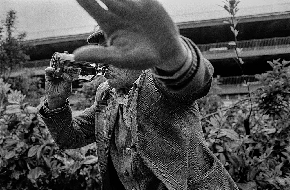 A man swigs from a bottle of spirits, waving his hand very close to the camera as if to hide his face.