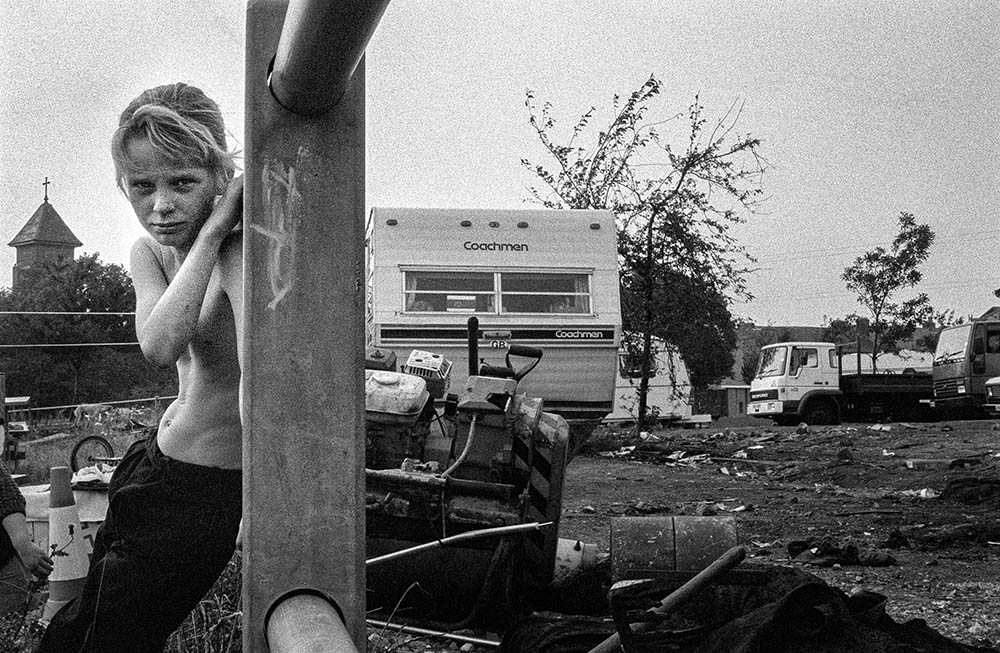 A child leans against a fence and frowns at the camera. Behind them is them is an area of wasteland with caravans and lorries parked up.