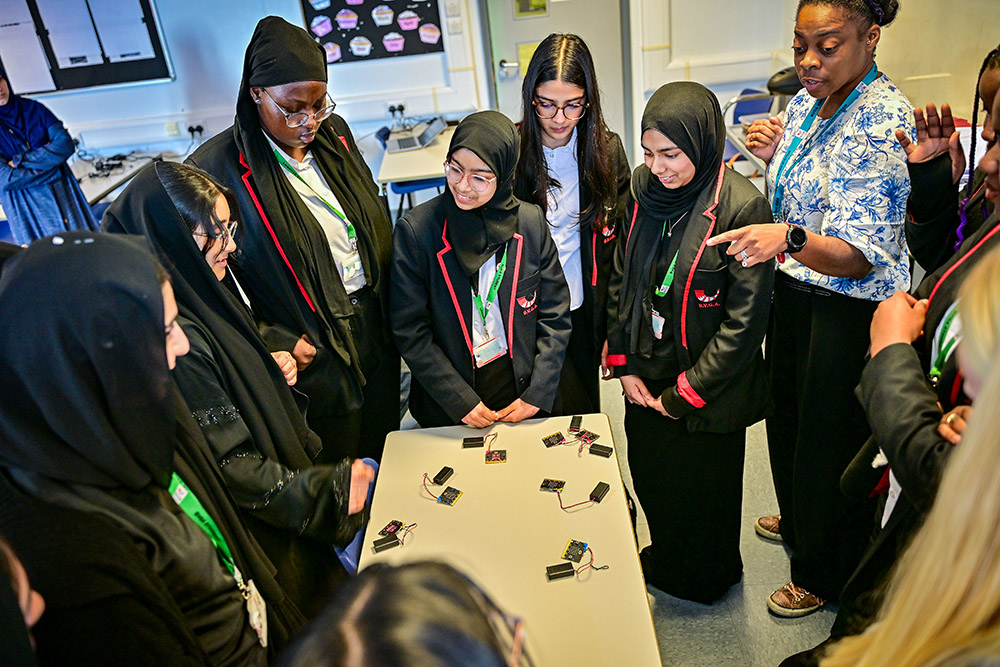 A group of students in uniform blazers stand around a table with small circuit boards.