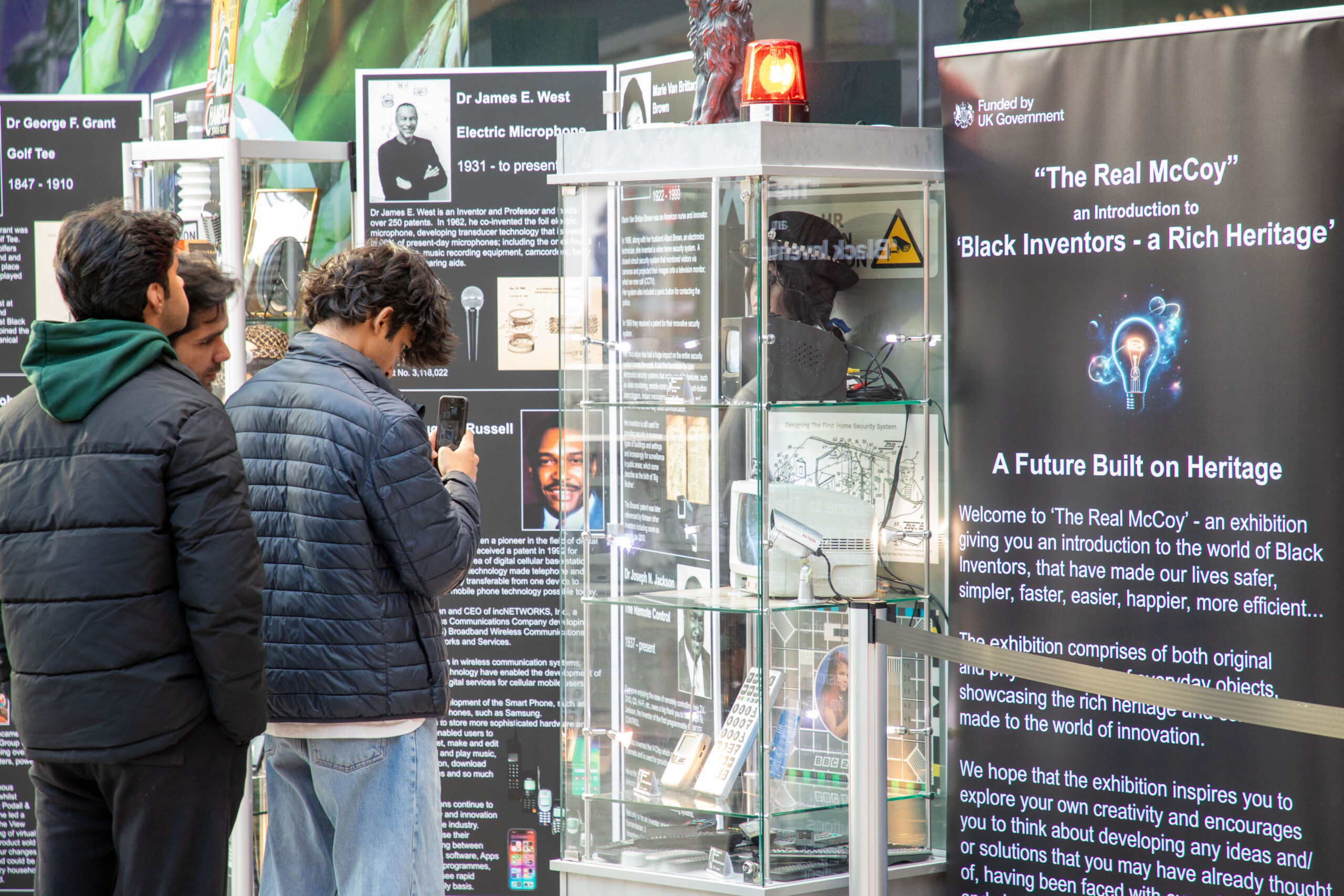 Three young men looking at the exhibition. One takes a picture on his mobile phone.