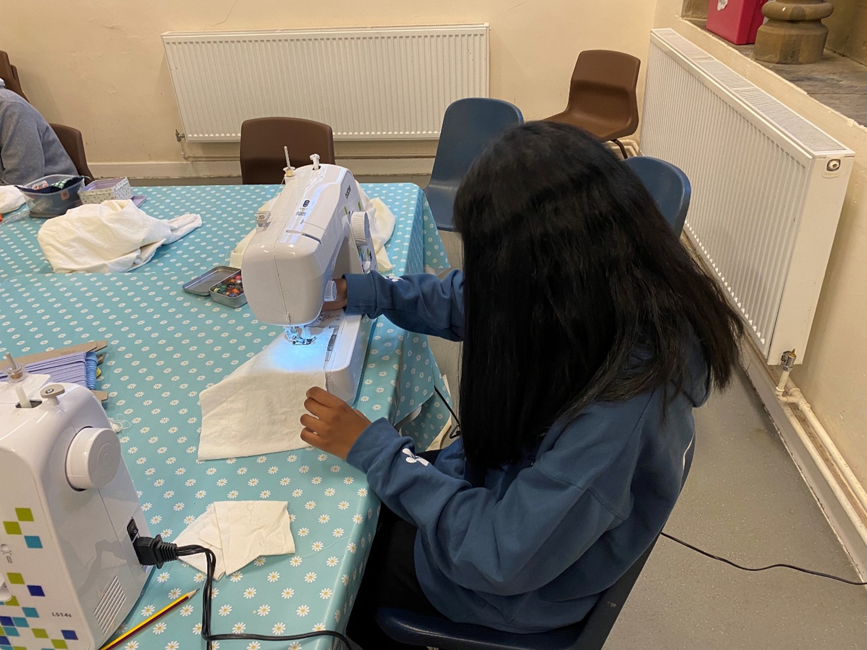 A teenager sitting at a sewing machine, sewing a small piece of white fabric