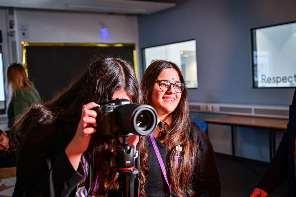 Two teenage girls smile as one looks through the viewfinder of a digital SLR on a tripod