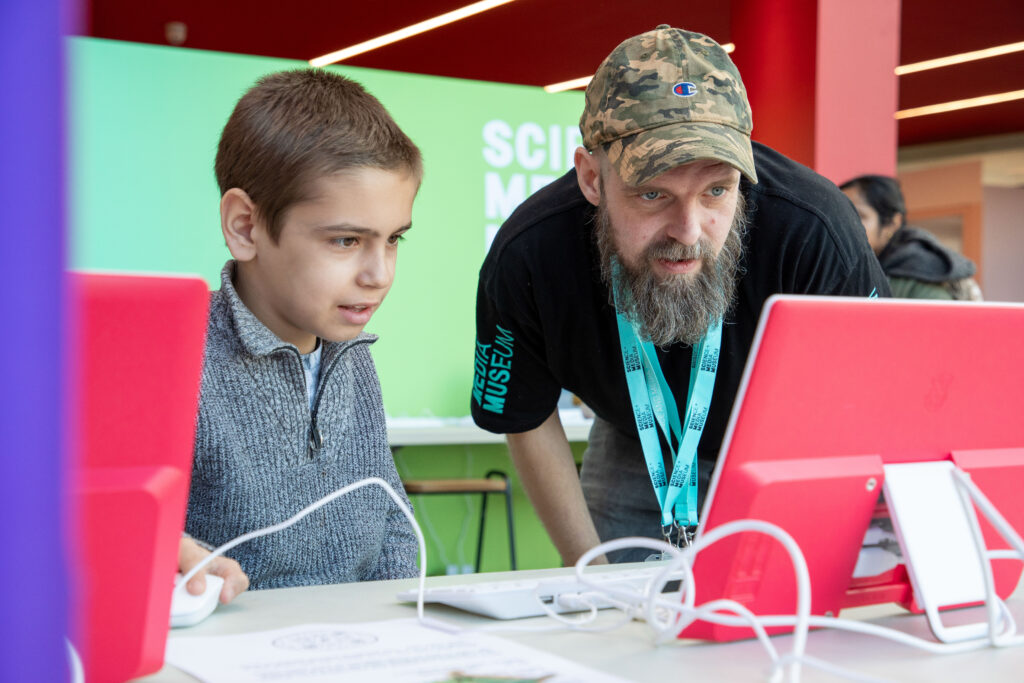 A man with a beard and baseball cap in Science and Media Museum volunteer uniform helps a young child with a computer game. They both look intently at the screen.