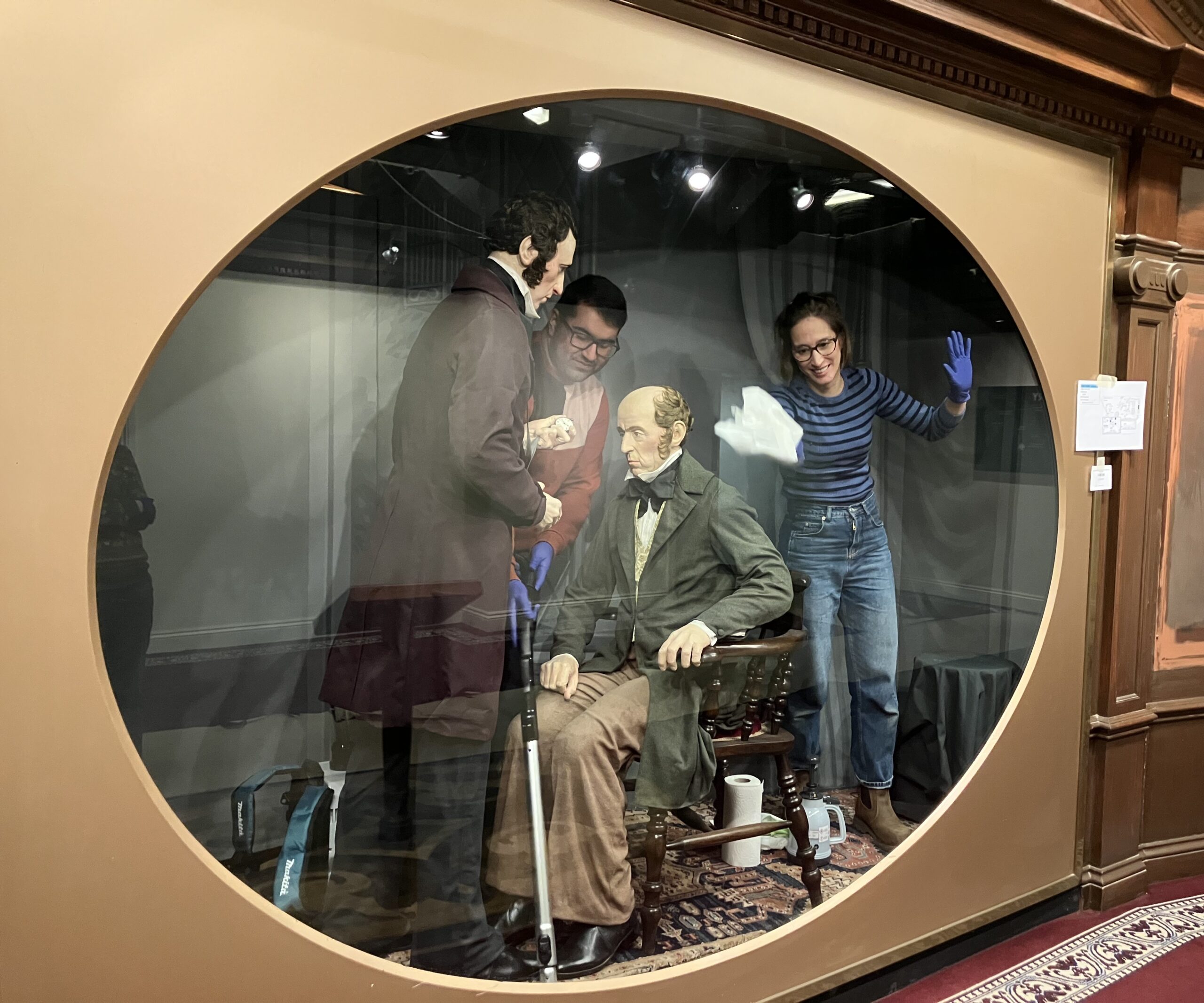 A glass museum case with two dummies of a Victorian photographer and his sitter. Saquib and Vanessa are behind the glass cleaning the display and smiling.