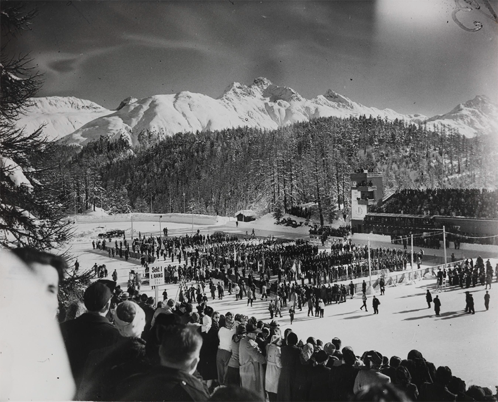 Crowds of people watch from bleachers as athletes gather in on a snowy showground with imposing snowy mountains in the background.