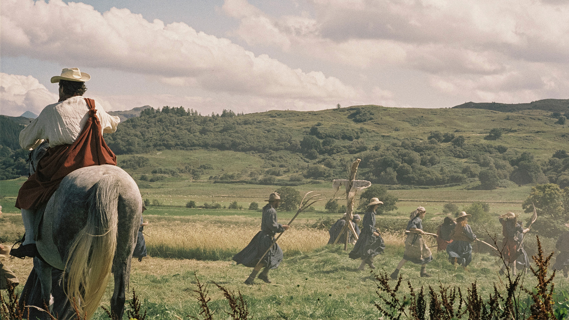 A man rides a grey horse away from the camera across an English field. A group of people with pitchforks and scythers walk in the background.