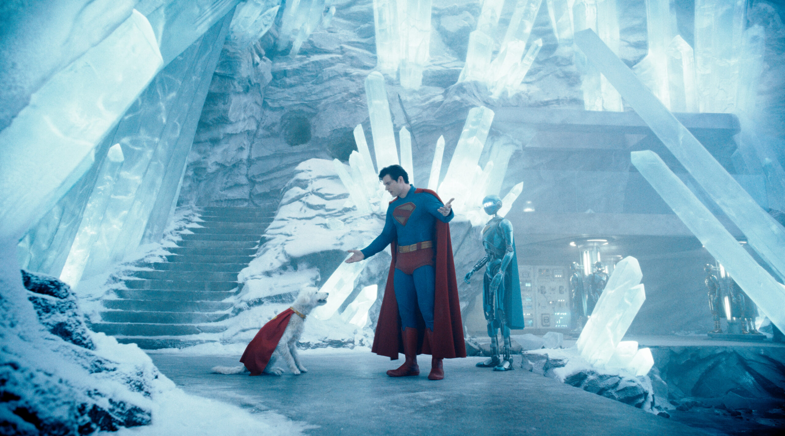 Superman stands in an icy cave, gesturing towards a white dog wearing a red cape. A robot stands behind him.