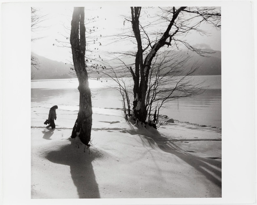 A woman walks with her head bowed through a serene lakeside landscape. She is slightly silhouetted by the low winter sun.