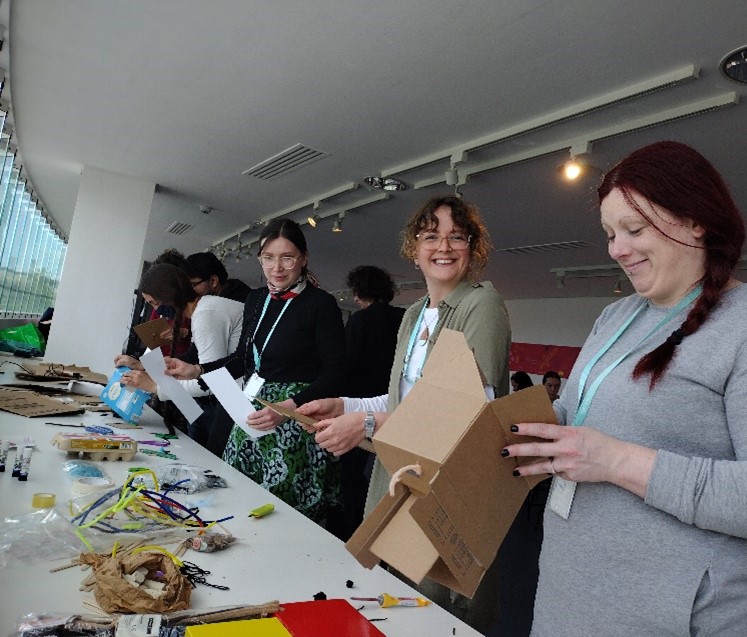Three people stand at a table covered with tinkering materials, looking excited