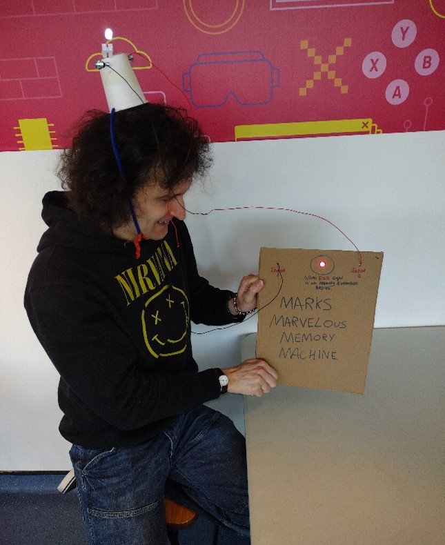 Person holds a cardboard model machine with wires connected to a small headpiece with a light