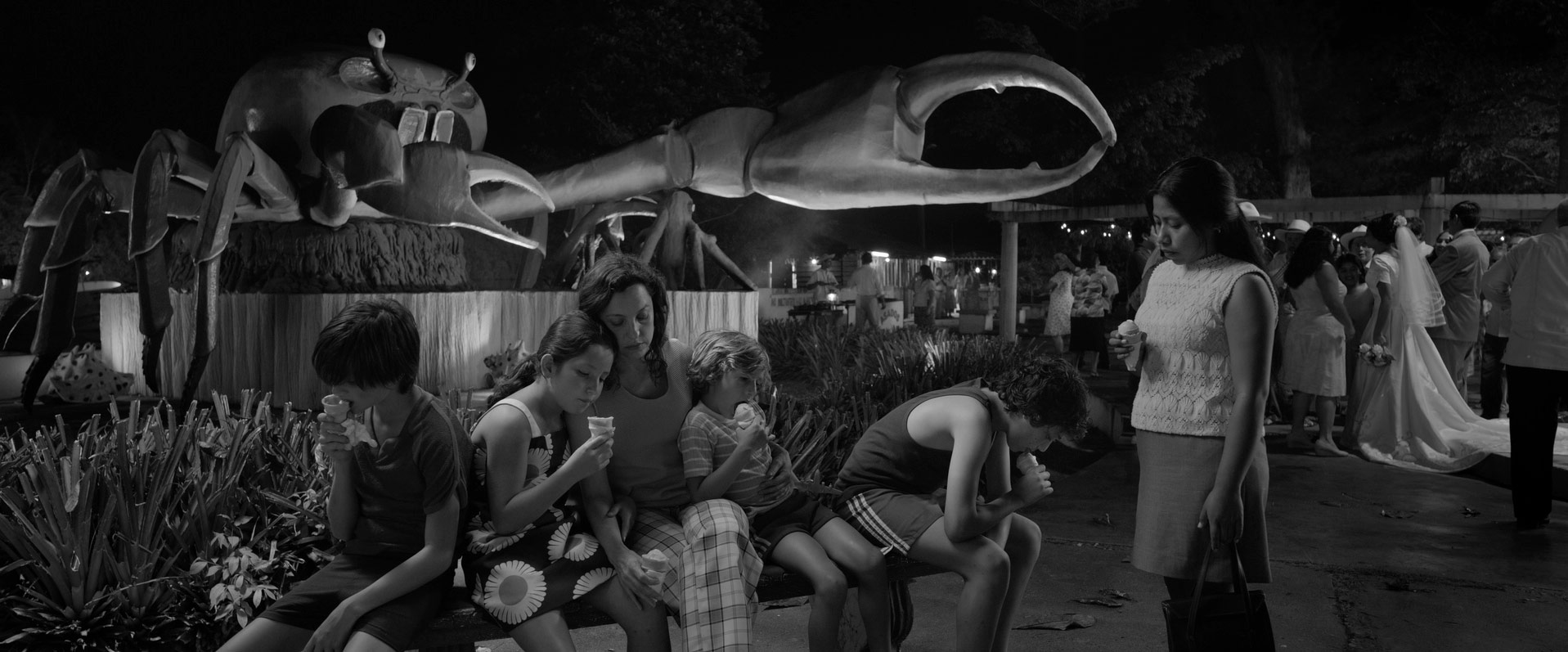 A group of women and children eat ice creams at a wedding, a large model crab looms behind them
