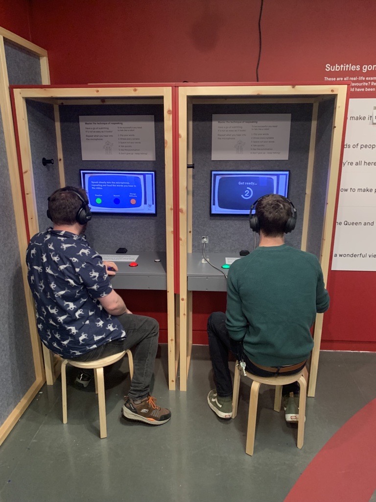 Two white men sit in booths with video screens wearing headphones