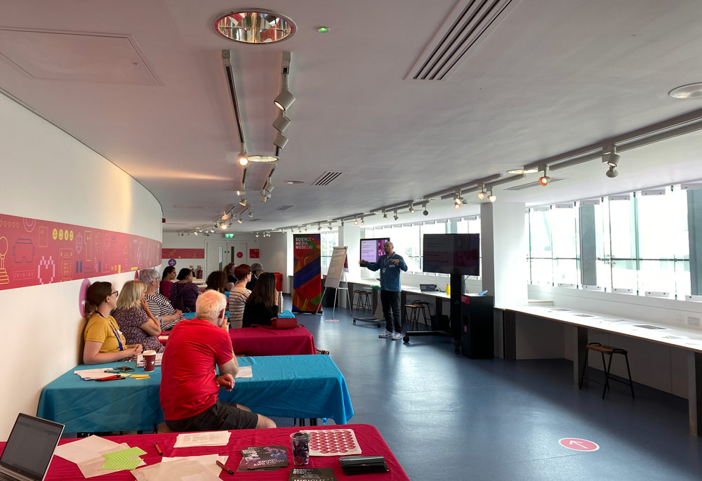 People sit at tables in a long, curved room listing to a man speak