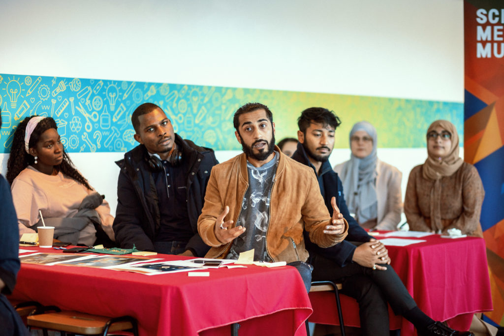 A group of people of various age, race and gender sit around small stables for a discussion.