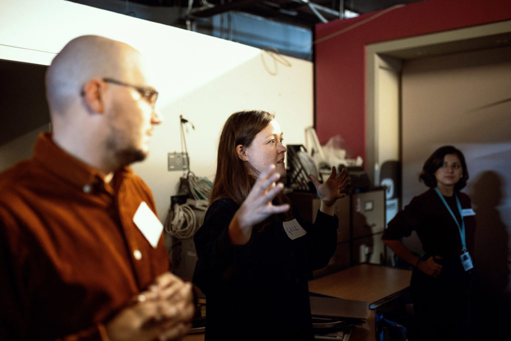 Three people stand in a dimly lit museum space; the woman in the centre gestures as she speaks