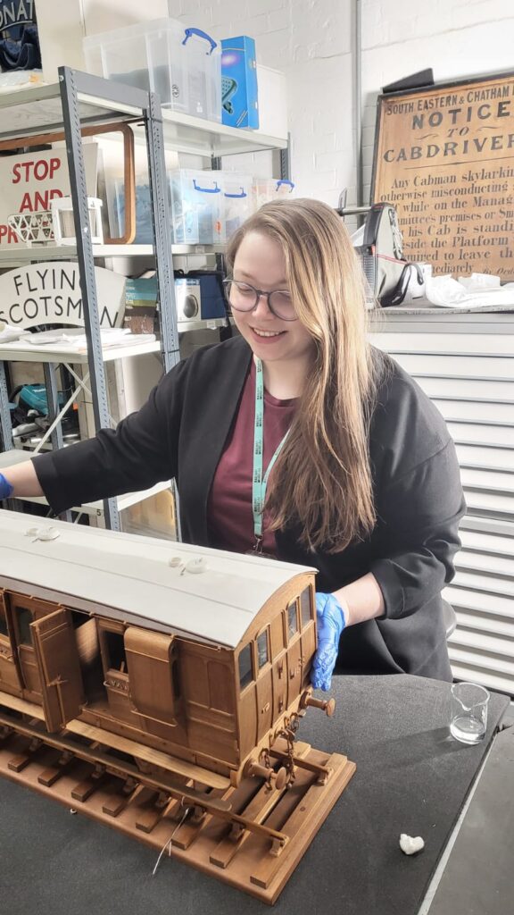 Emily, a white woman with long hair and glasses, handles a model train carriage in the stores