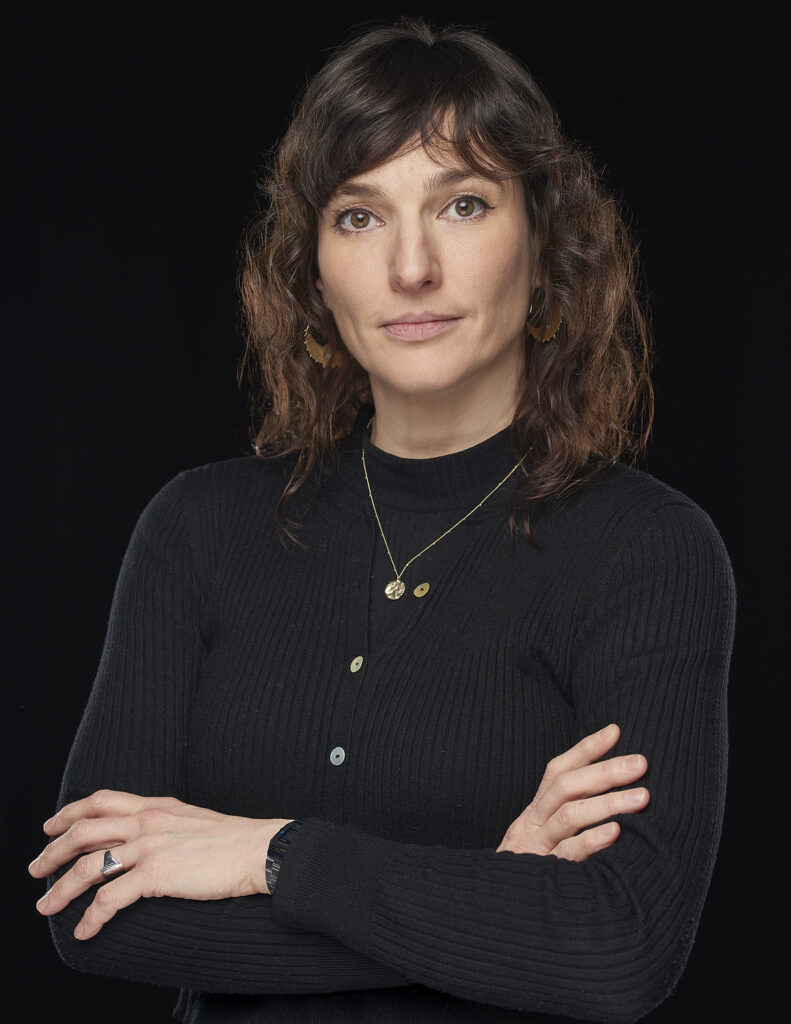 Lucy is a white woman with curly brown hair wearing all black. She looks pensively at the camera.