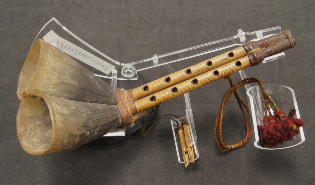 A pipe-shaped musical instrument with a colourful tassel, mounted on a clear support