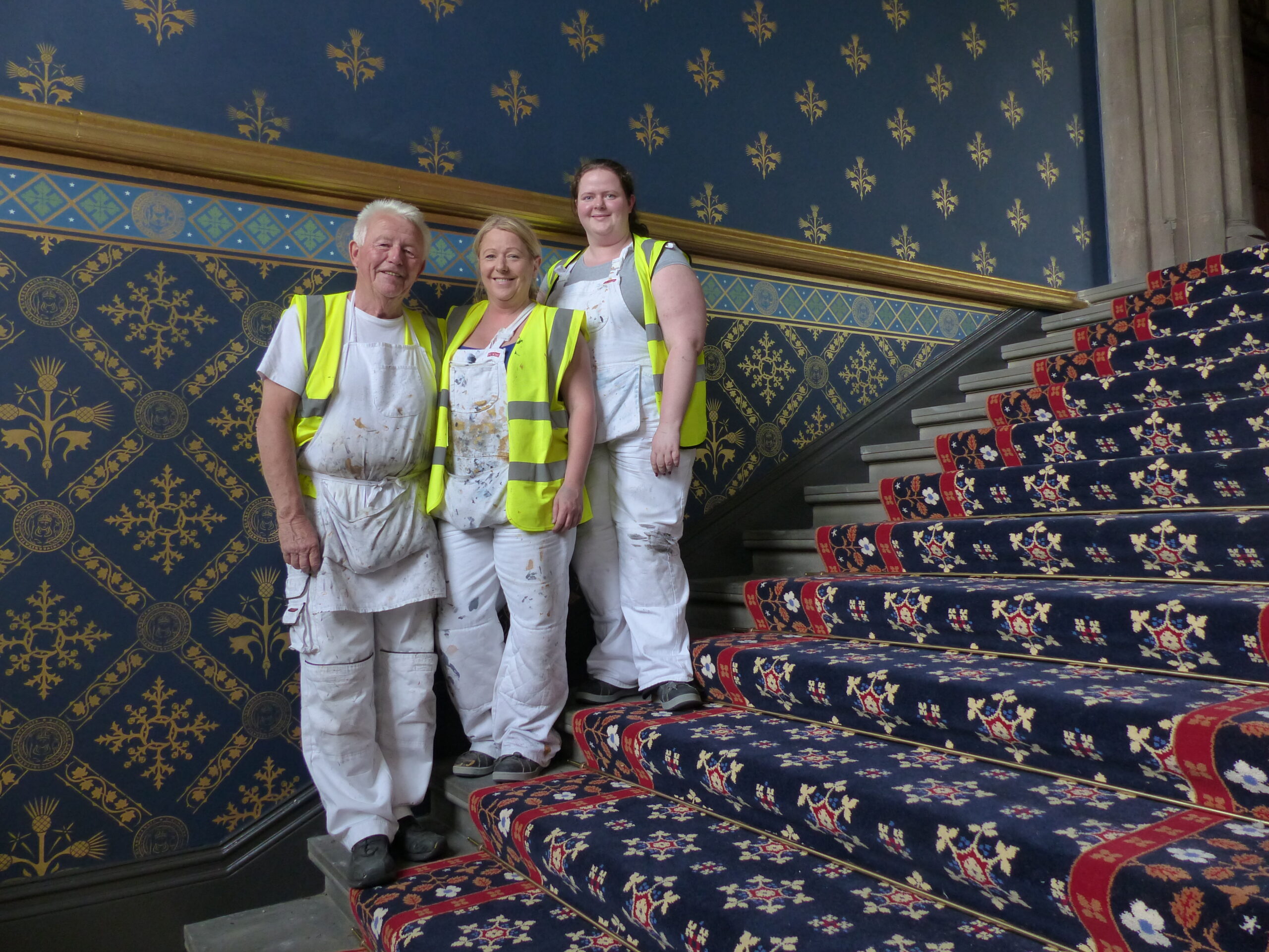 Rebecca stands on grand carpeted stairs wtih two other people, all wearing white overalls