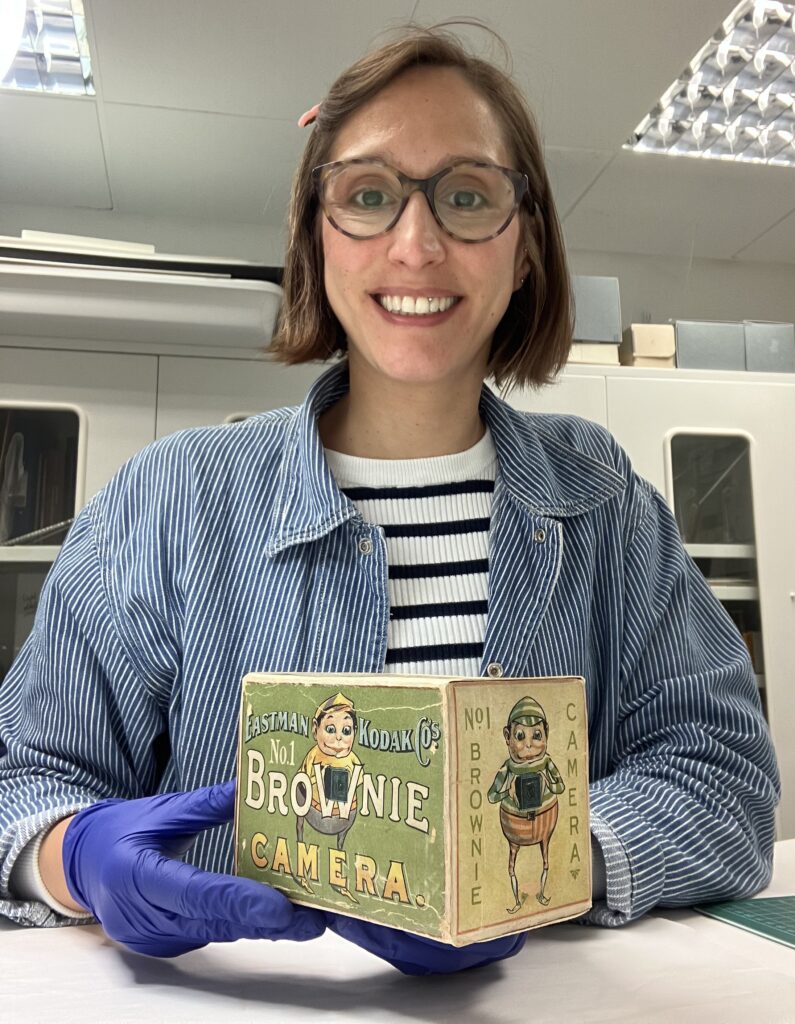 Vanessa smiles to the camera, wearing blue nitrile gloves and holding the Brownie box