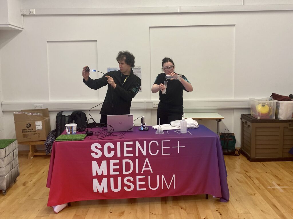 Two explainers doing a science experiement at a table with a Science + Media Museum banner