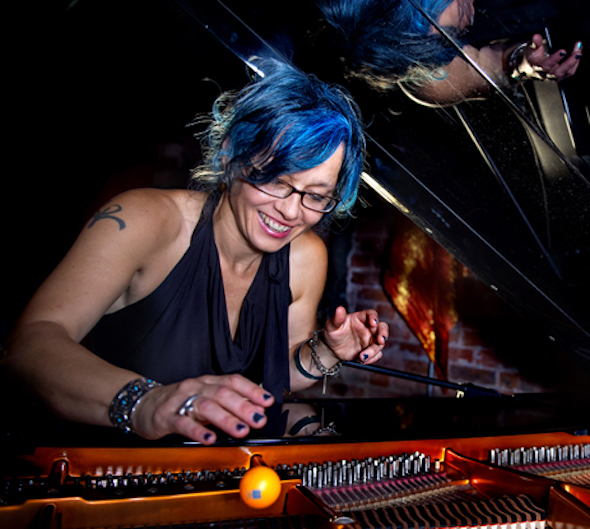 Caro looks inside a grand piano, smiling. She is white, with blue hair and glasses.