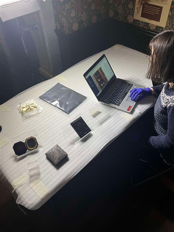Ruth, a white woman, sits at a table of museum objects, wearing blue nitrile gloves and using a laptop.